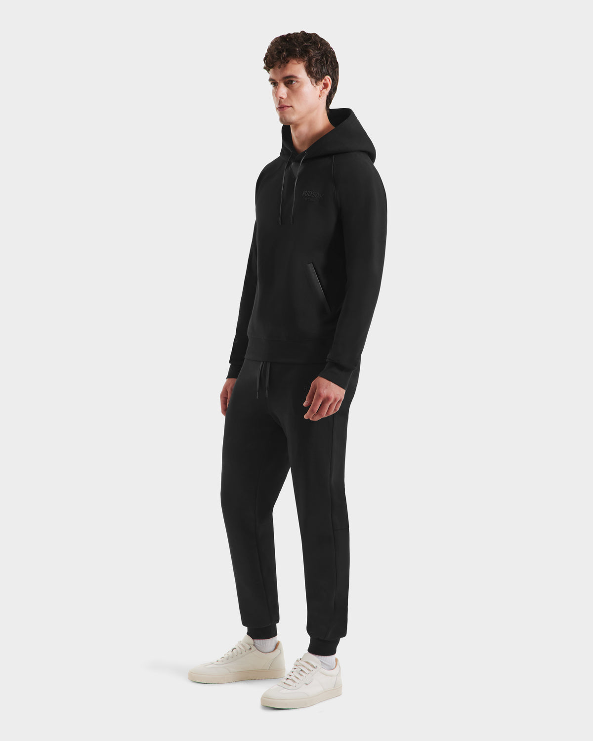 A man models the Rudsak BROOKLYN - 6525593 Black hoodie against a plain white background, paired with matching joggers and white sneakers for a modern urban look. He stands slightly turned, hands relaxed at his sides.
