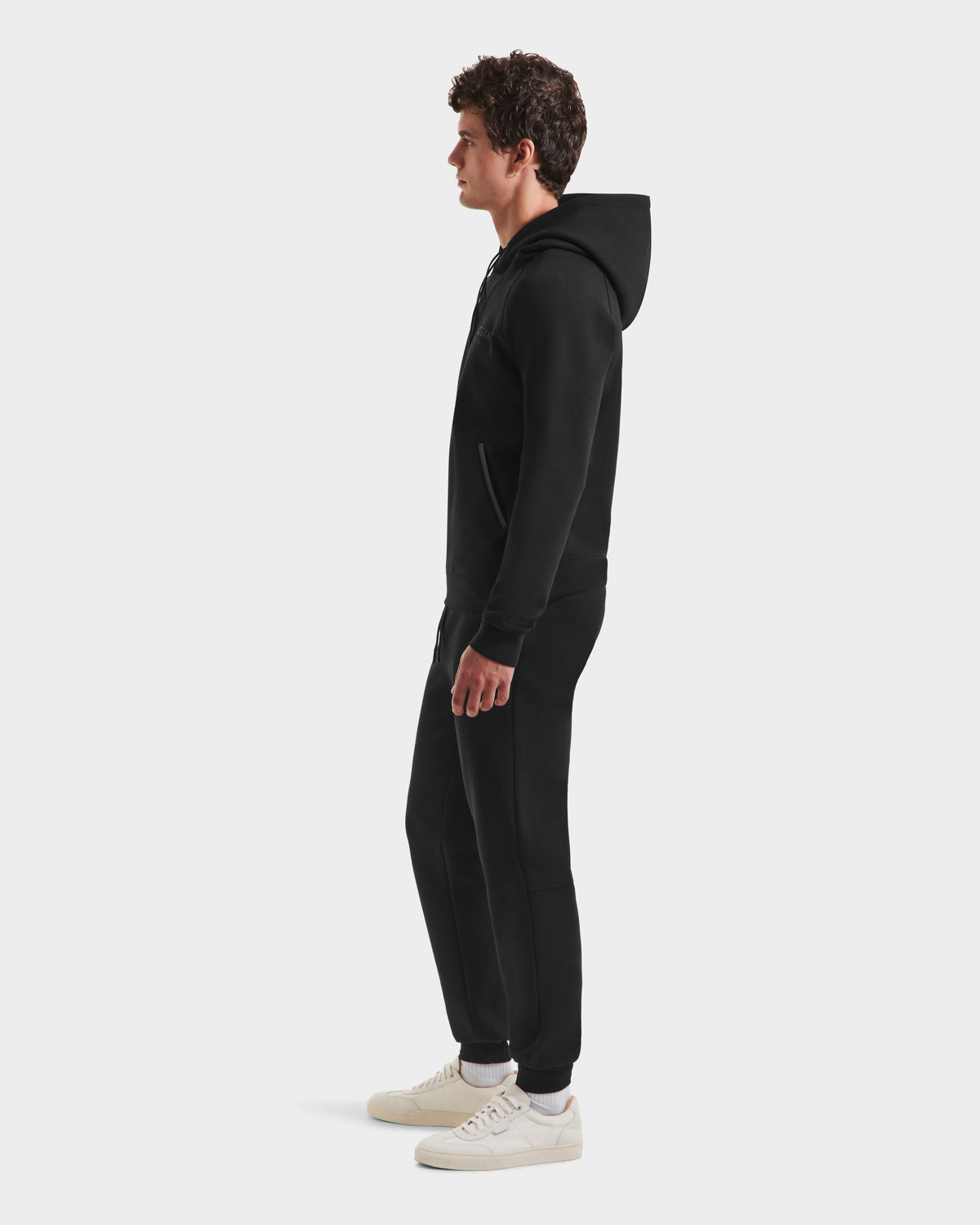 A man in profile faces left, wearing the Rudsak BROOKLYN - 6525593 Black hoodie and joggers with white sneakers, showcasing urban style against a plain white background.