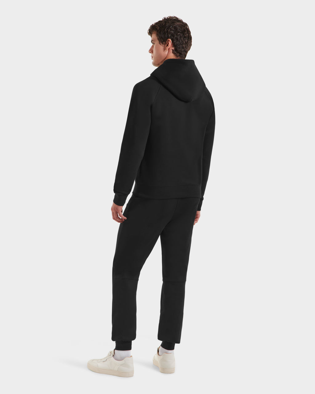 A person stands facing away, showcasing urban style in Rudsak’s BROOKLYN - 6525593 Black men’s hoodie and black pants with white sneakers against a plain white background.
