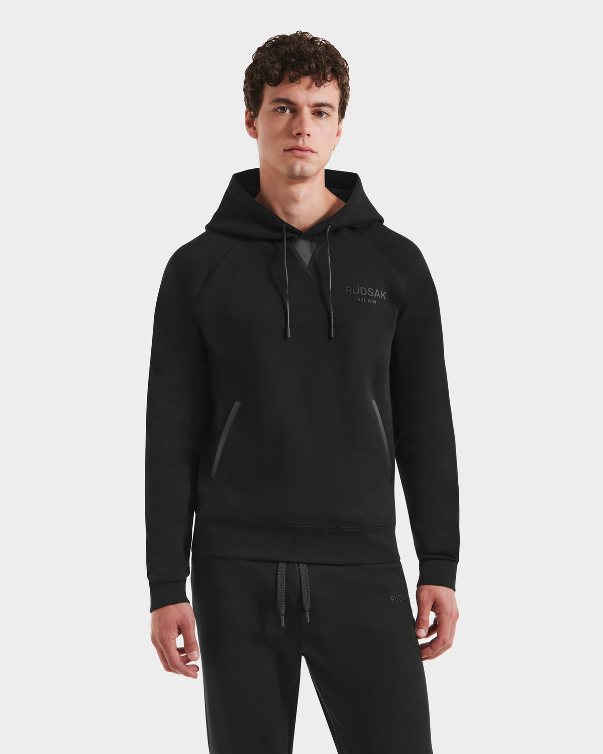 A young man with curly brown hair wears the Rudsak BROOKLYN - 6525593 Black men’s hoodie and matching black sweatpants, standing forward with his hands at his sides against a plain white background.
