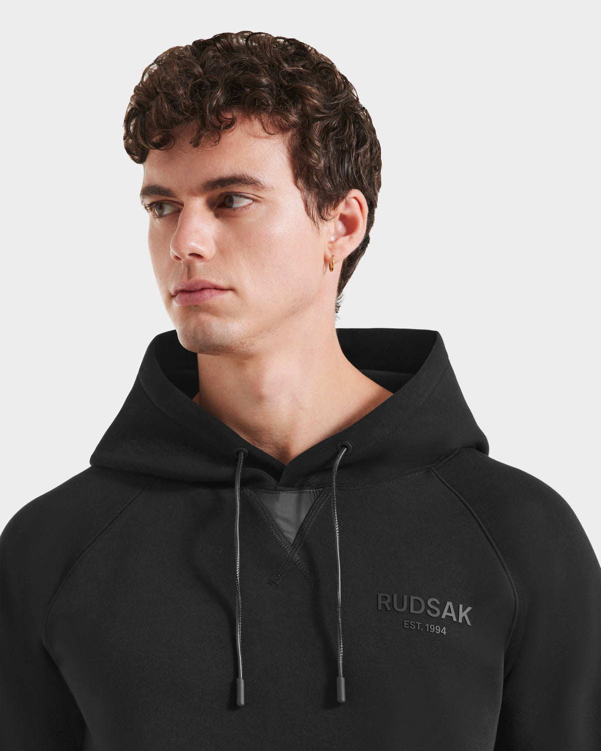 A young man with curly brown hair models the Rudsak BROOKLYN - 6525593 Black hoodie featuring RUDSAK EST. 1994 print, embodying urban style as he looks to his left against a plain white background.