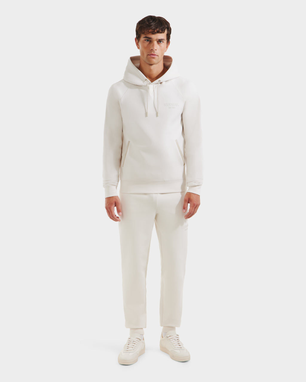 A man faces forward against a white background, wearing the Rudsak BROOKLYN - 6525593 Vanilla hoodie with matching jogger pants and sneakers.