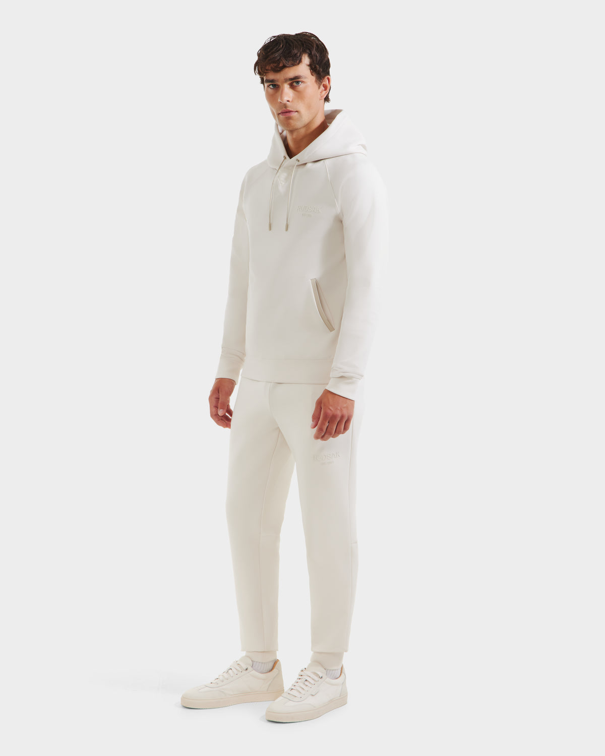 A man stands against a white background, wearing the Rudsak BROOKLYN - 6525593 Vanilla cotton-blend hoodie and matching jogger pants, paired with light sneakers. He faces the camera with a neutral expression.