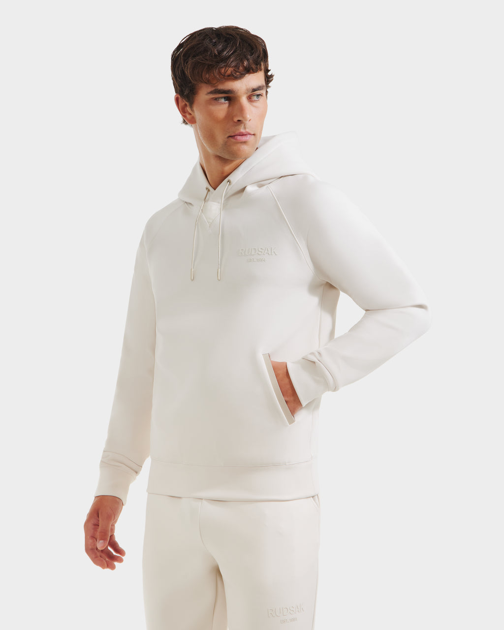 A man wears the Rudsak BROOKLYN - 6525593 Vanilla cotton-blend hoodie and matching sweatpants, right hand in the pocket, looking left against a plain white background.