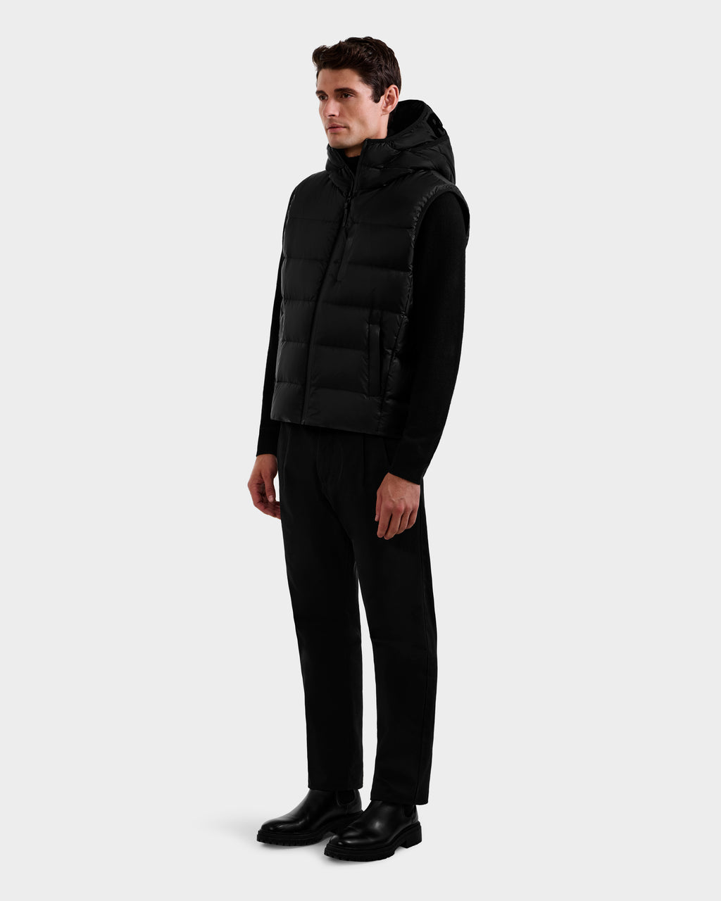A man stands against a plain background wearing the Rudsak BROOKS - M125520 Black men’s down puffer vest over a black long-sleeve shirt, black pants, and black shoes. He faces slightly right with arms relaxed at his sides.