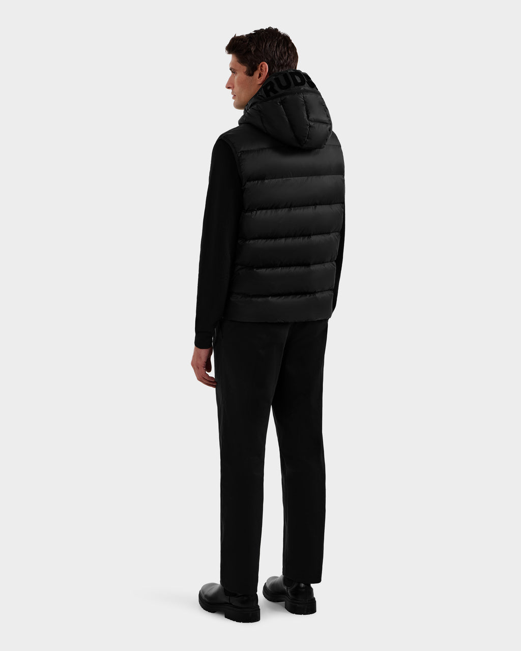 A man faces away in a Rudsak BROOKS - M125520 Black hooded down vest with a recycled-nylon shell, layered over a black long-sleeve top, black pants, and black shoes, set against a light gray background.