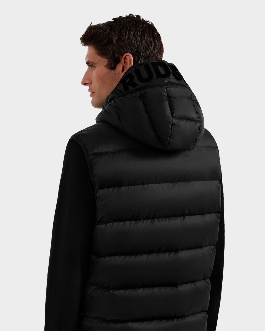A man with short brown hair is seen from the back wearing the Rudsak BROOKS - M125520 Black puffer jacket, featuring a hood crafted from recycled-nylon shell with bold black lettering. The background is plain and light-colored.