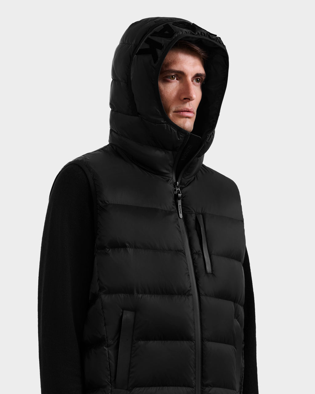 A person wears the Rudsak BROOKS - M125520 Black hooded down vest made from recycled-nylon shell, zipped up with the hood on, looking to the side against a plain light background.