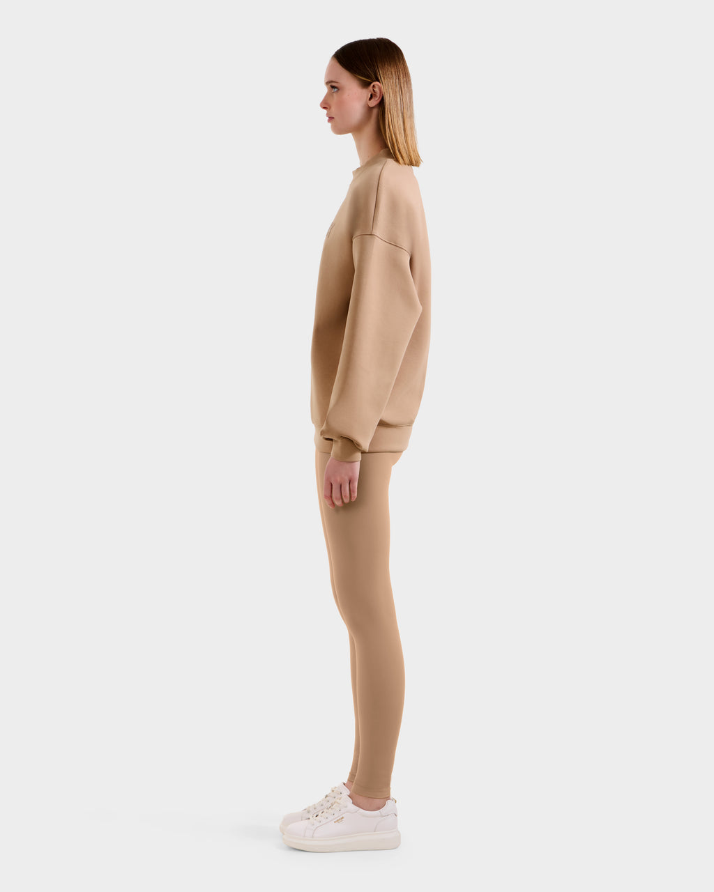 A woman stands in profile against a plain background, wearing the Rudsak BRYN - 9524007 Almond W beige cotton crewneck sweatshirt with genderless sizing, paired with matching beige leggings and white sneakers.