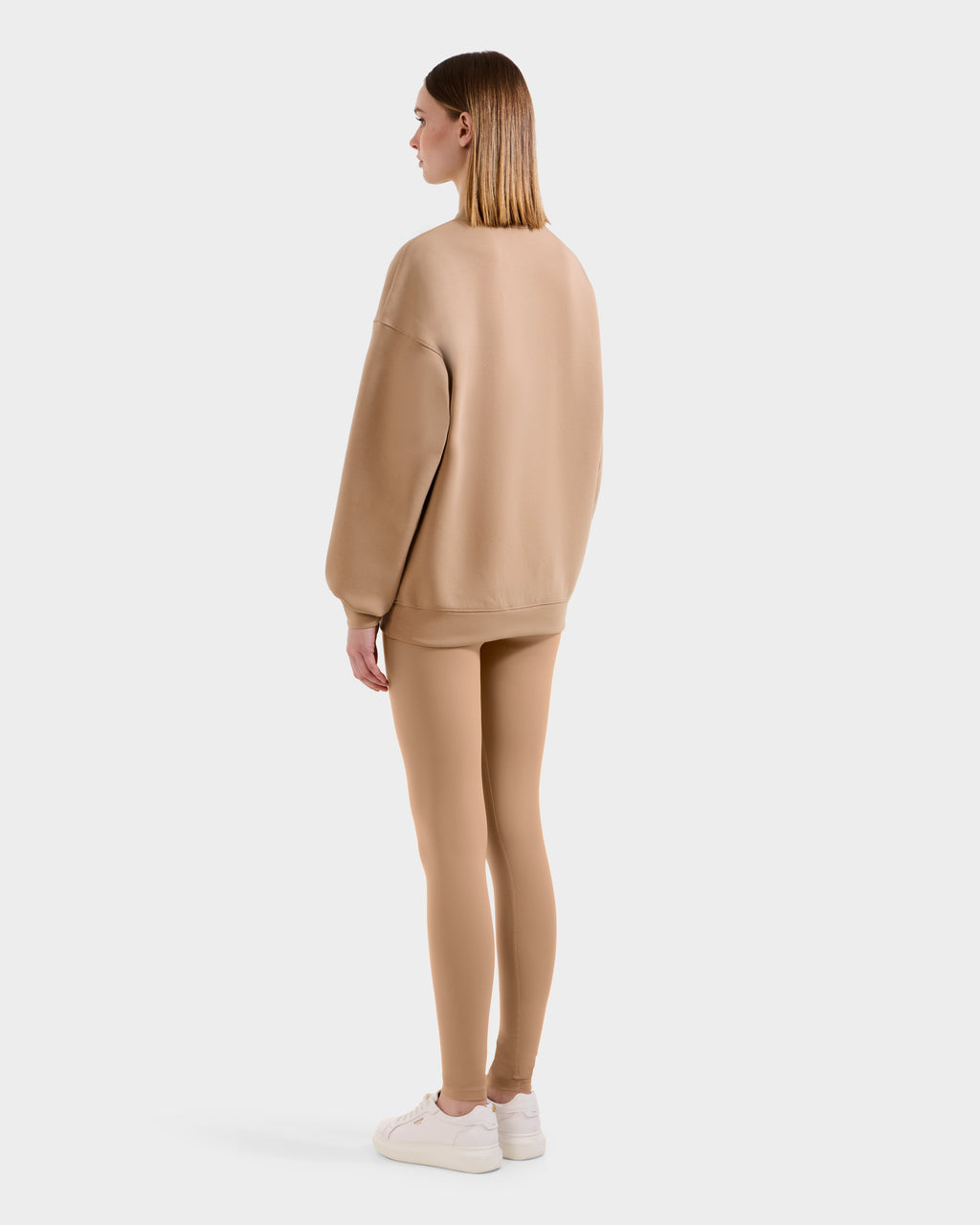 A woman with straight, shoulder-length hair faces away, wearing the Rudsak BRYN - 9524007 Almond W: a loose cotton crewneck sweatshirt with matching tan leggings and white sneakers—a perfect everyday fit against a plain white background.