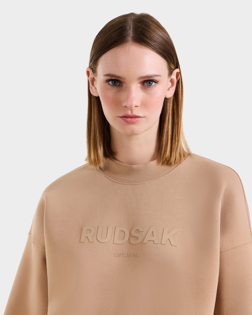 A woman with straight, shoulder-length light brown hair wears the Rudsak BRYN - 9524007 Almond W beige cotton crewneck sweatshirt with "RUDSAK EST. 1994" embossed on the front, standing against a plain light gray background.