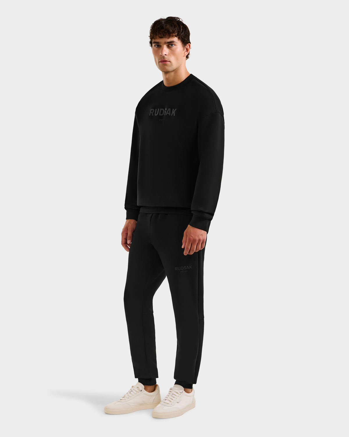 A man stands facing left in a Rudsak BRYN - 9524007 Black M crewneck sweatshirt and matching joggers, both in everyday fit and genderless sizing, paired with white sneakers against a light gray background.