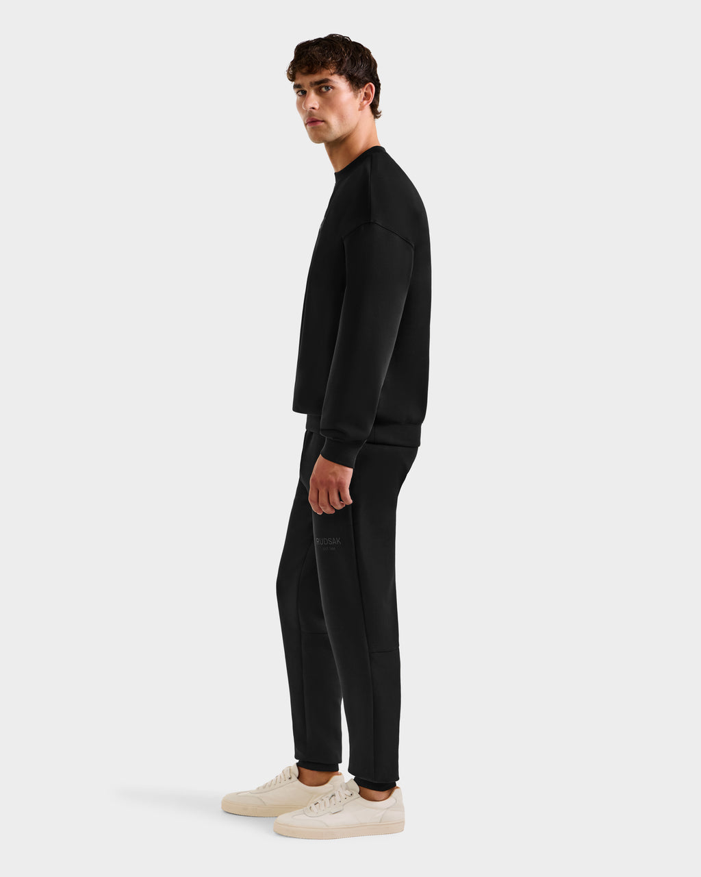 A man stands sideways, looking ahead, wearing the Rudsak BRYN - 9524007 Black M cotton crewneck sweatshirt and matching pants with white sneakers, presenting a stylish everyday look against a plain white background.
