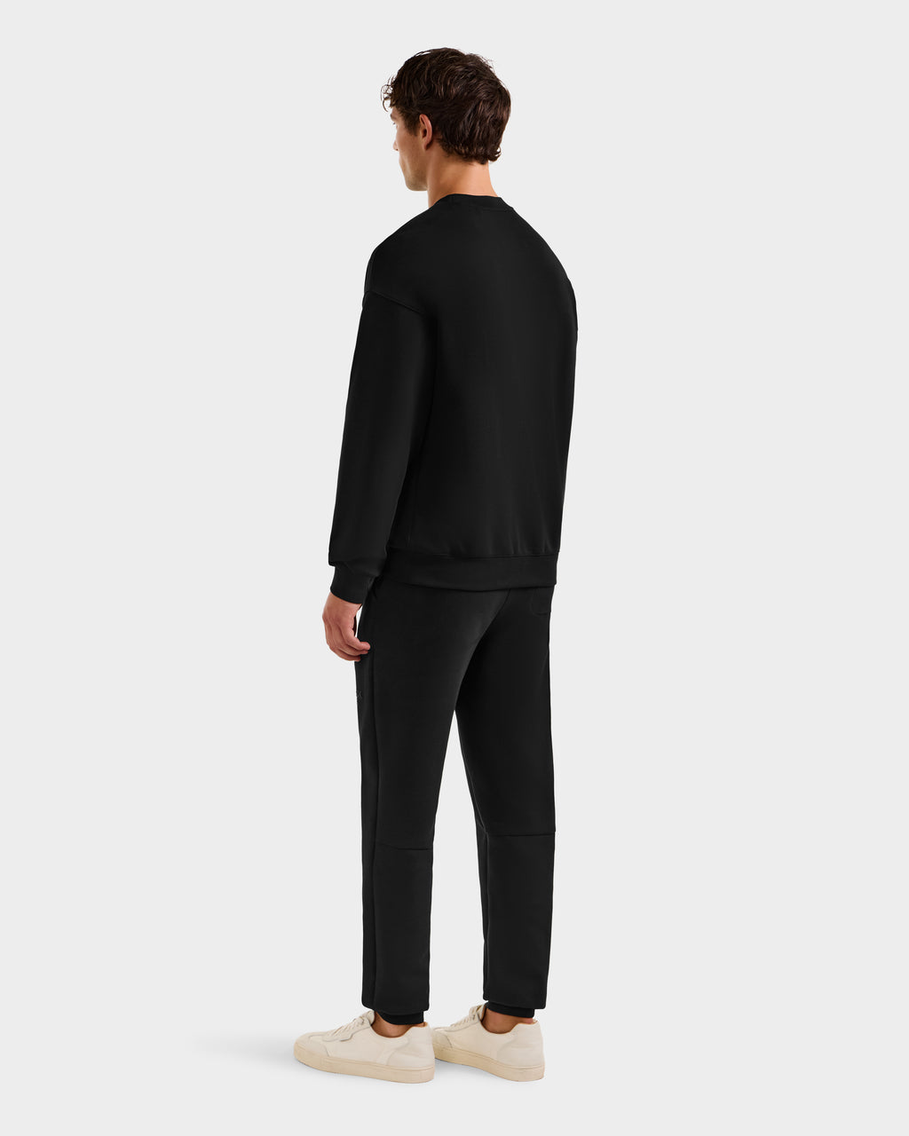 A man faces away, wearing the Rudsak BRYN - 9524007 Black M cotton crewneck sweatshirt and jogger pants with white sneakers, displaying a genderless fit against a plain white background.