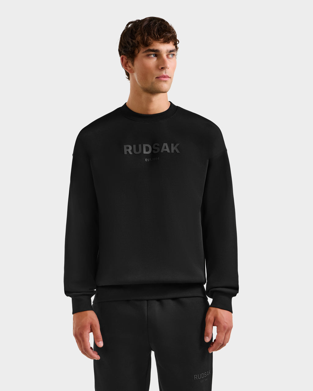 A young man with short brown hair wears the Rudsak BRYN - 9524007 black cotton crewneck sweatshirt in size M, featuring “RUDSAK” on the chest and a genderless, everyday fit. He pairs it with black pants and stands against a light gray background.