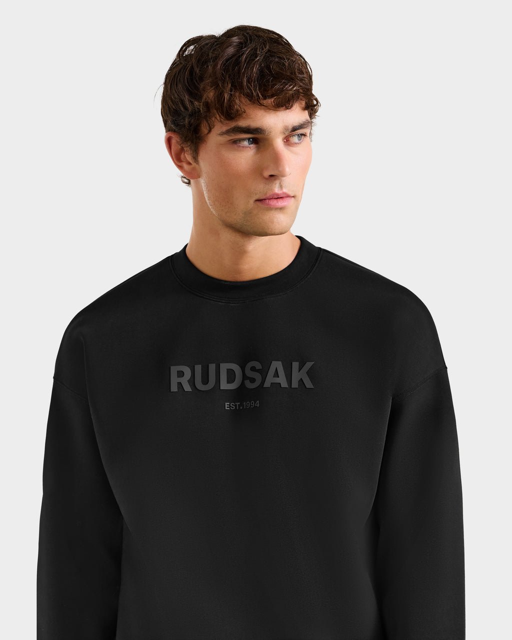 A young man with short brown hair wears the Rudsak BRYN - 9524007 Black M cotton crewneck sweatshirt, featuring RUDSAK and EST. 1994 on the chest. He looks to the side against a plain light background.
