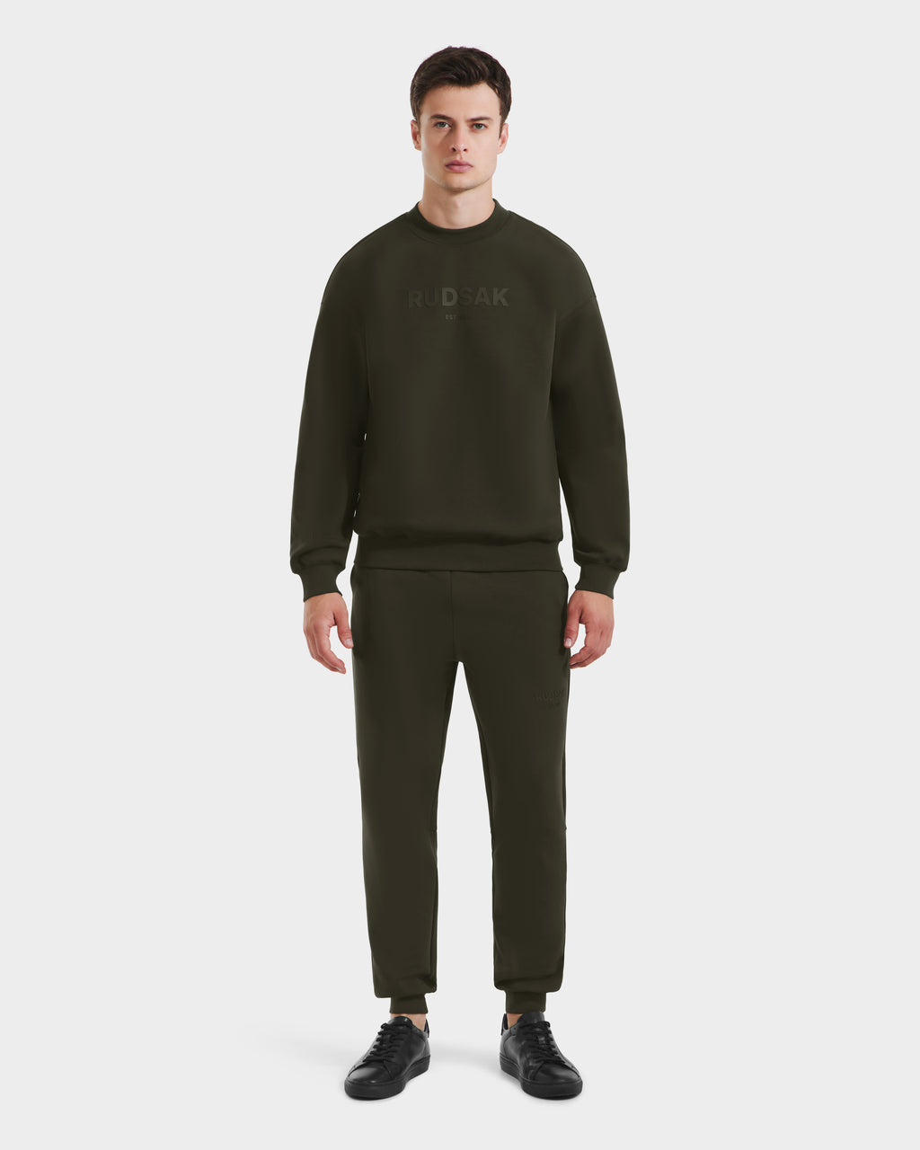 A man faces forward in a plain white setting, wearing the Rudsak BRYN - 9524007 Dark Olive M crewneck sweatshirt and matching joggers, paired with black sneakers.