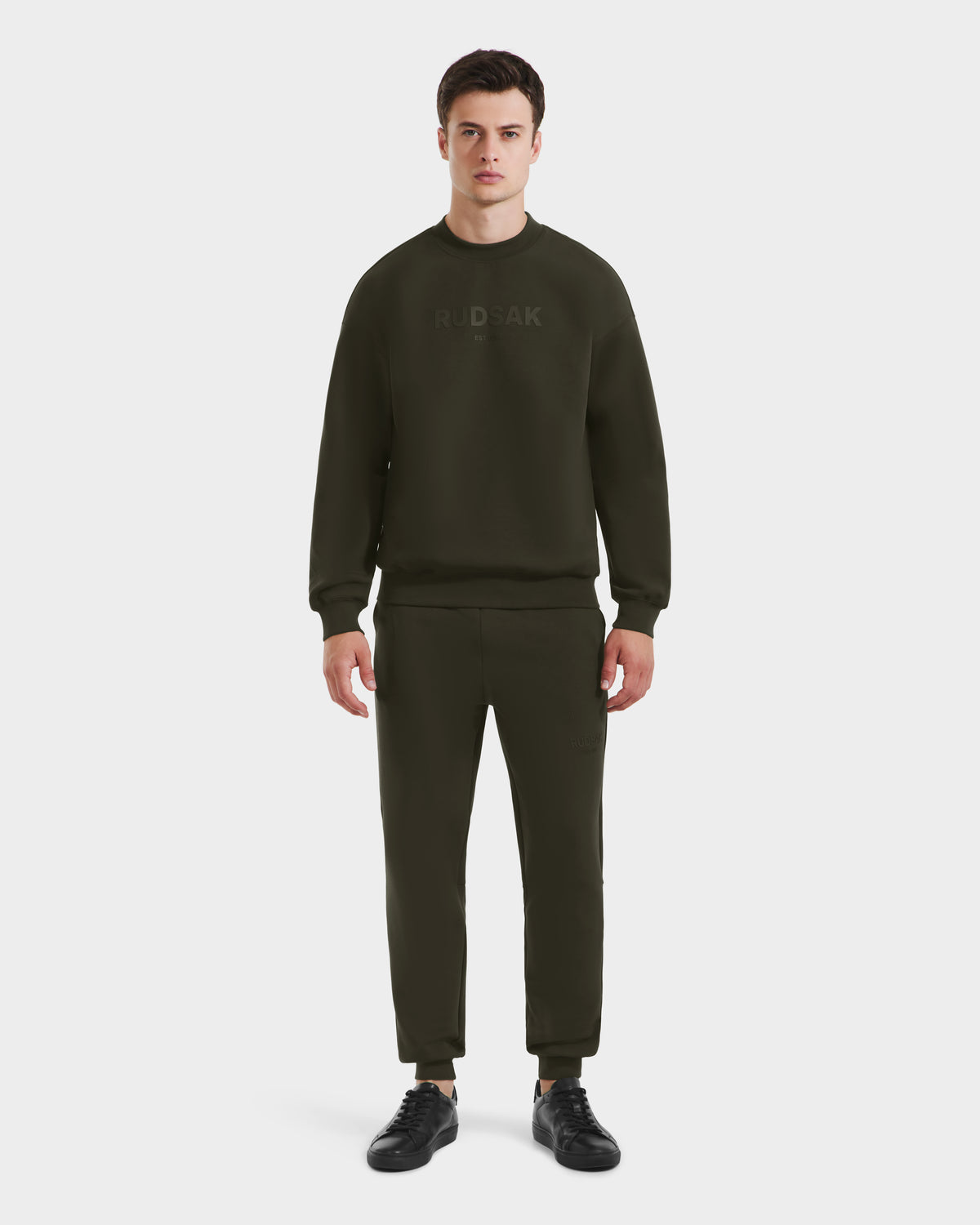 A man faces forward in a plain white setting, wearing the Rudsak BRYN - 9524007 Dark Olive M crewneck sweatshirt and matching joggers, paired with black sneakers.
