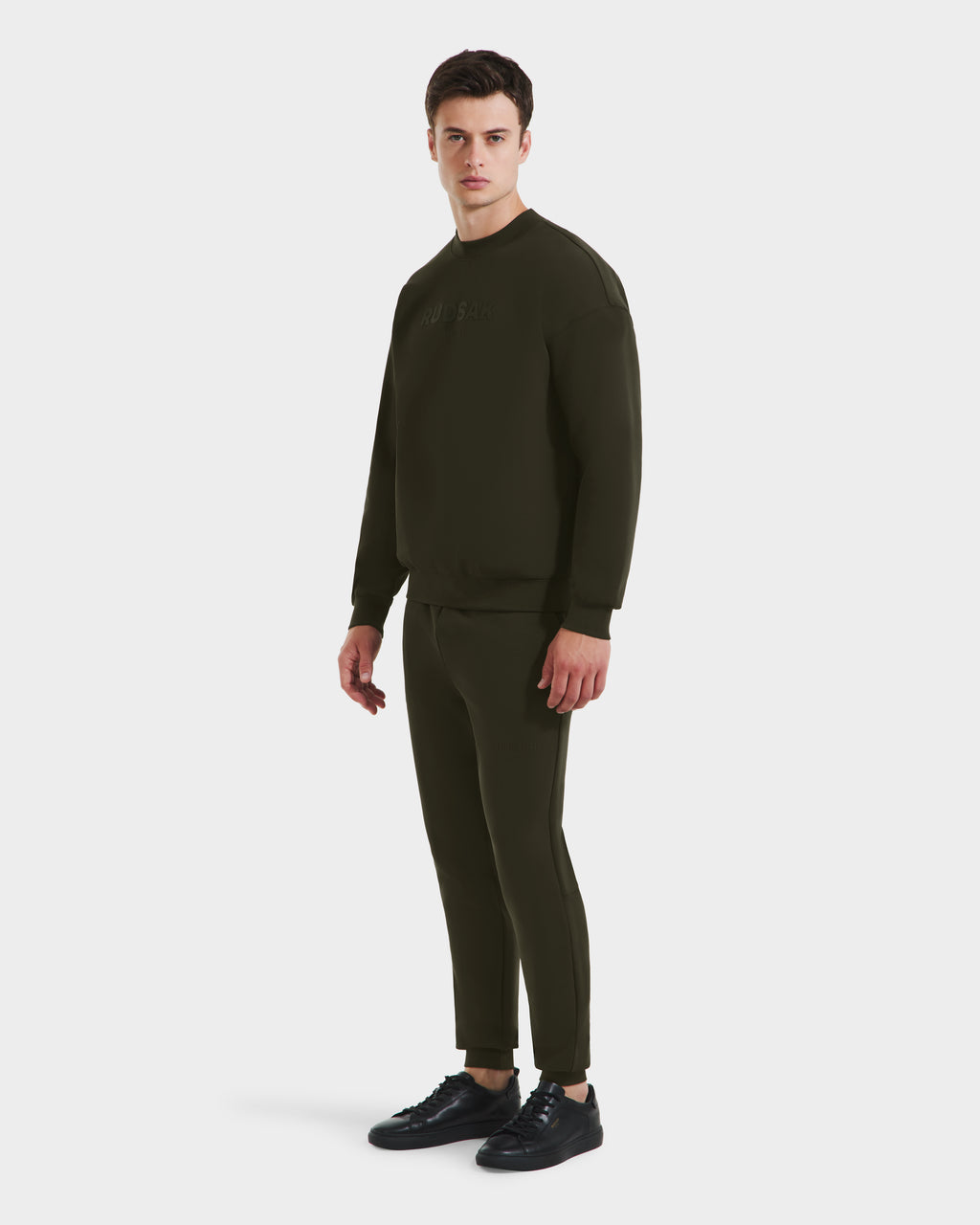 A man stands slightly sideways in the Rudsak BRYN - 9524007 Dark Olive M cotton crewneck sweatshirt and jogger set with black sneakers, on a white background—an everyday fit featuring genderless sizing.