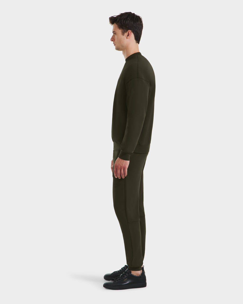 A man stands in profile wearing the Rudsak BRYN - 9524007 Dark Olive M sweatshirt and joggers set with black sneakers, highlighting everyday style against a plain white background.