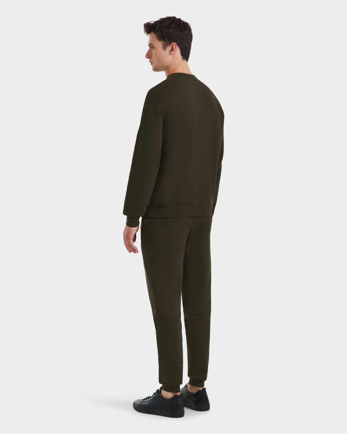 A man models the Rudsak BRYN - 9524007 Dark Olive M cotton crewneck sweatshirt and pants set with black sneakers—an easy, genderless everyday look—against a plain white background.