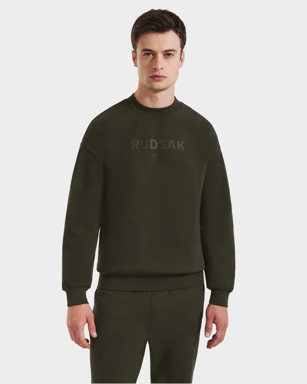 A man stands against a white background wearing the Rudsak BRYN - 9524007 crewneck sweatshirt and matching sweatpants in Dark Olive, featuring subtle front branding—an ideal choice for everyday style.