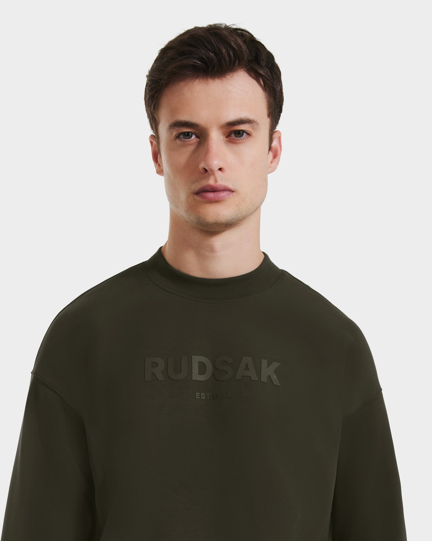 A young man with short brown hair wears the Rudsak BRYN - 9524007 crewneck sweatshirt in dark olive, featuring an embossed logo, everyday fit, and genderless sizing, standing against a plain white background.