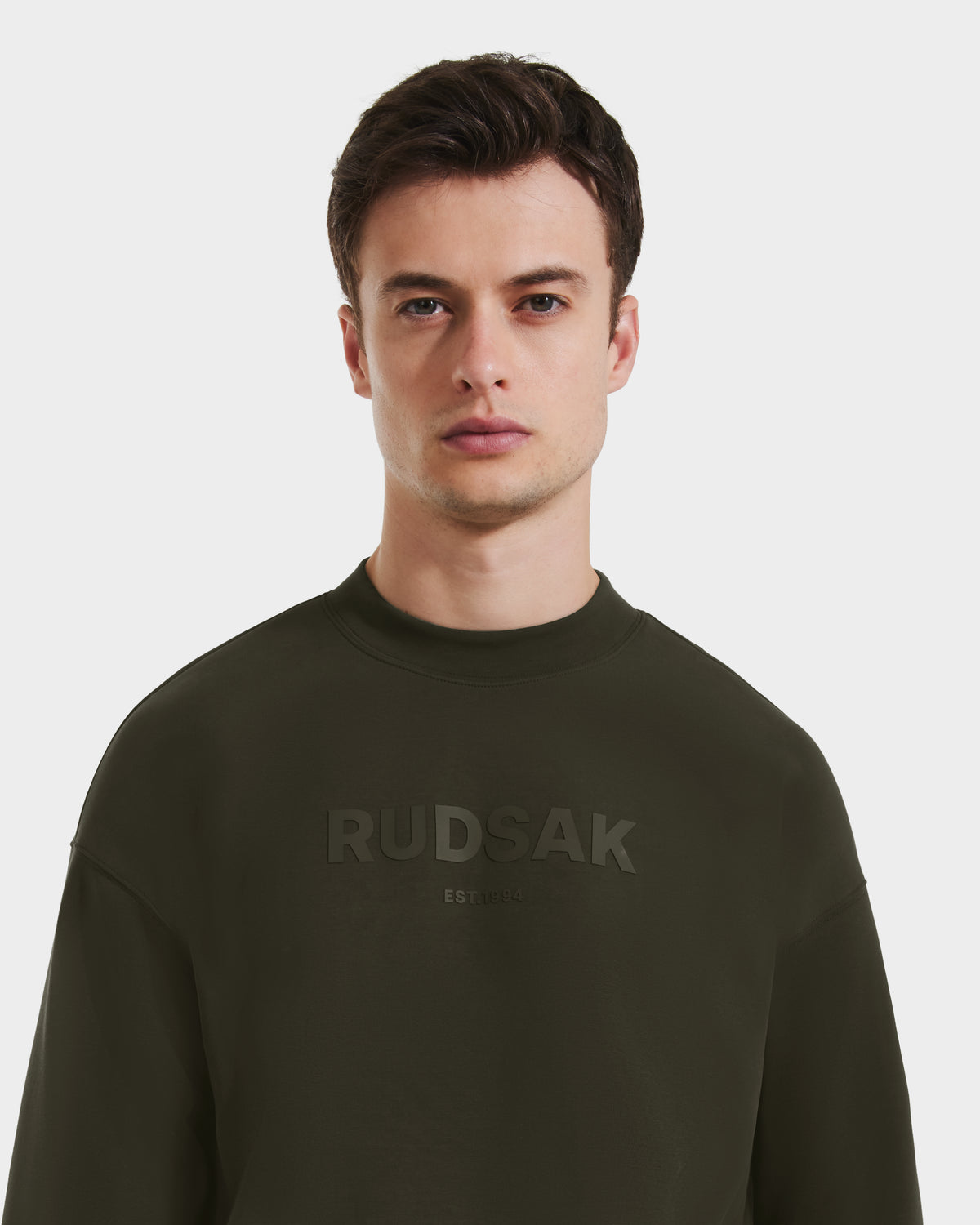 A young man with short brown hair wears the Rudsak BRYN - 9524007 crewneck sweatshirt in dark olive, featuring an embossed logo, everyday fit, and genderless sizing, standing against a plain white background.