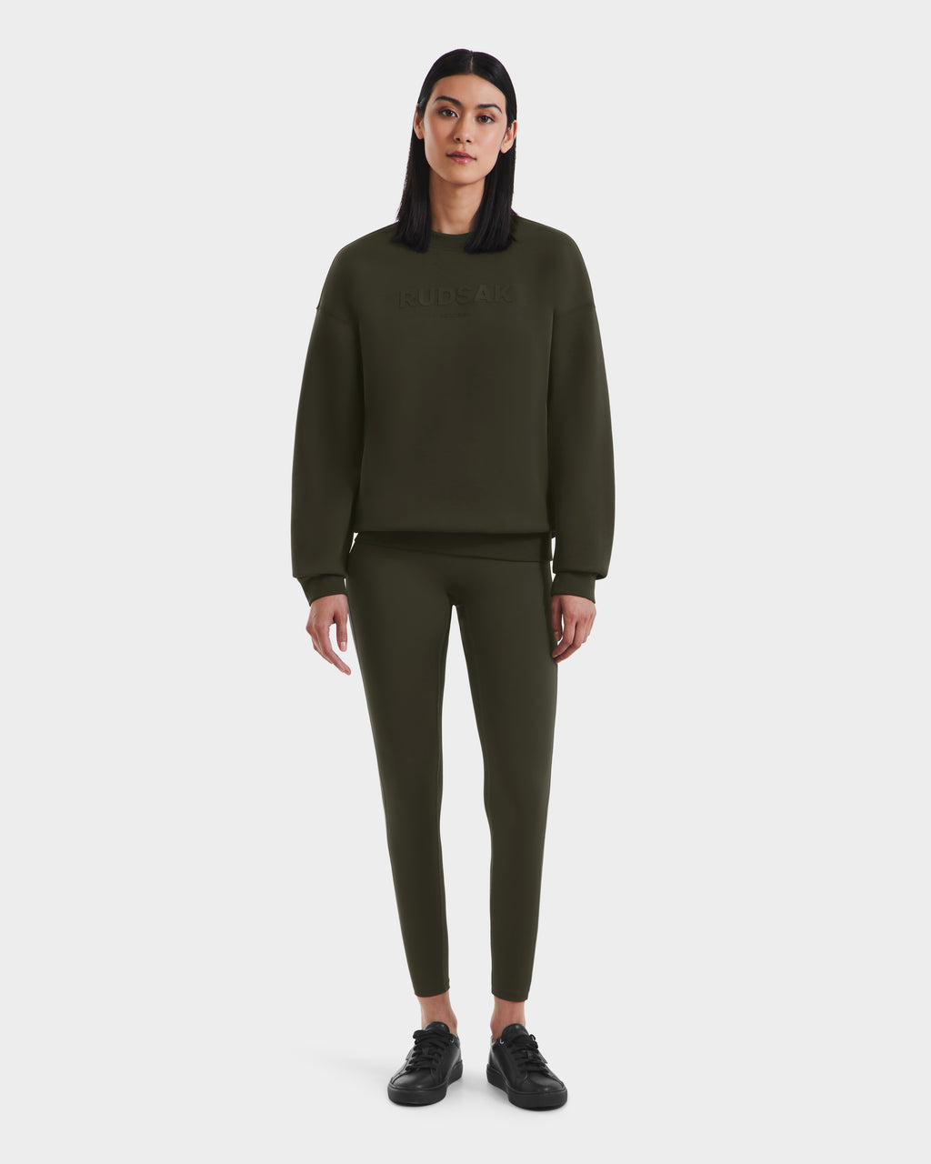 A woman with straight dark hair wears the Rudsak BRYN - 9524007 Dark Olive W cotton crewneck sweatshirt and matching fitted pants, paired with black sneakers, standing against a white background with a neutral expression—a timeless look.
