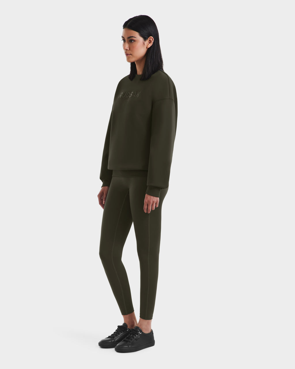 A woman with straight dark hair wears the Rudsak BRYN - 9524007 Dark Olive W cotton crewneck sweatshirt and matching leggings, styled with black sneakers, highlighting timeless design and genderless sizing on a white background.