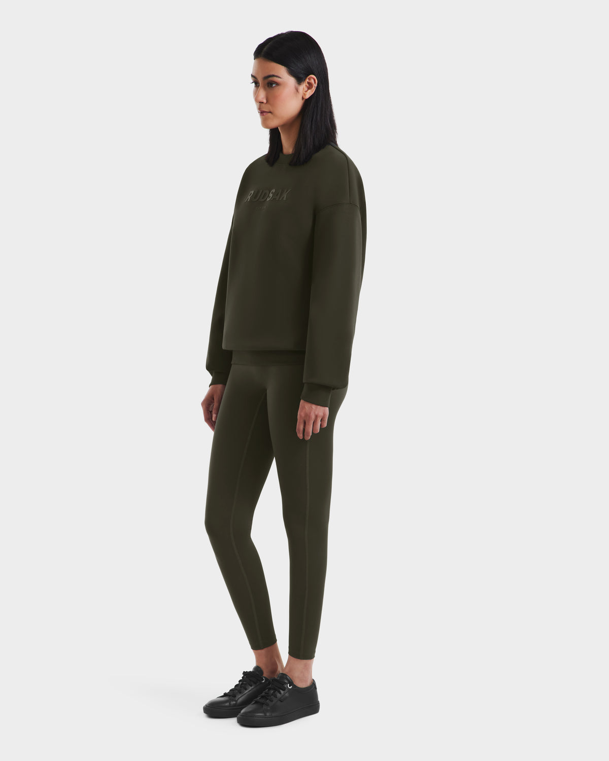 A woman with straight dark hair wears the Rudsak BRYN - 9524007 Dark Olive W cotton crewneck sweatshirt and matching leggings, styled with black sneakers, highlighting timeless design and genderless sizing on a white background.