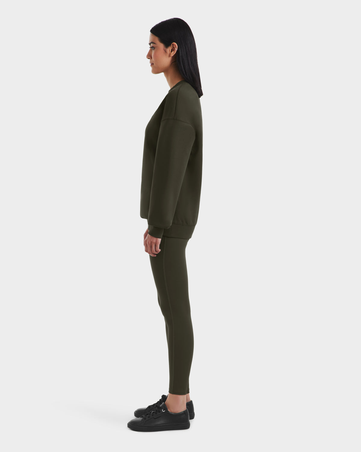 A woman stands in profile against a white background, wearing the Rudsak BRYN - 9524007 Dark Olive W cotton crewneck sweatshirt and matching leggings with black sneakers—timeless pieces in genderless sizing for versatile wear.