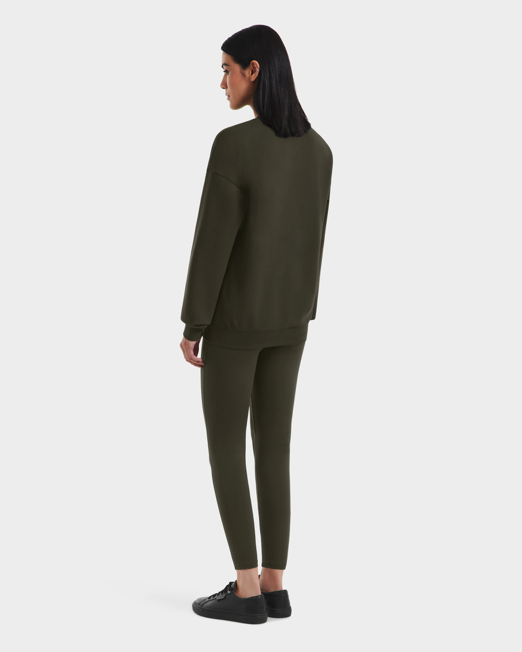 A person with straight dark hair faces away, wearing Rudsaks BRYN - 9524007 Dark Olive W cotton crewneck sweatshirt and matching leggings in genderless sizing, paired with black sneakers against a plain white background.