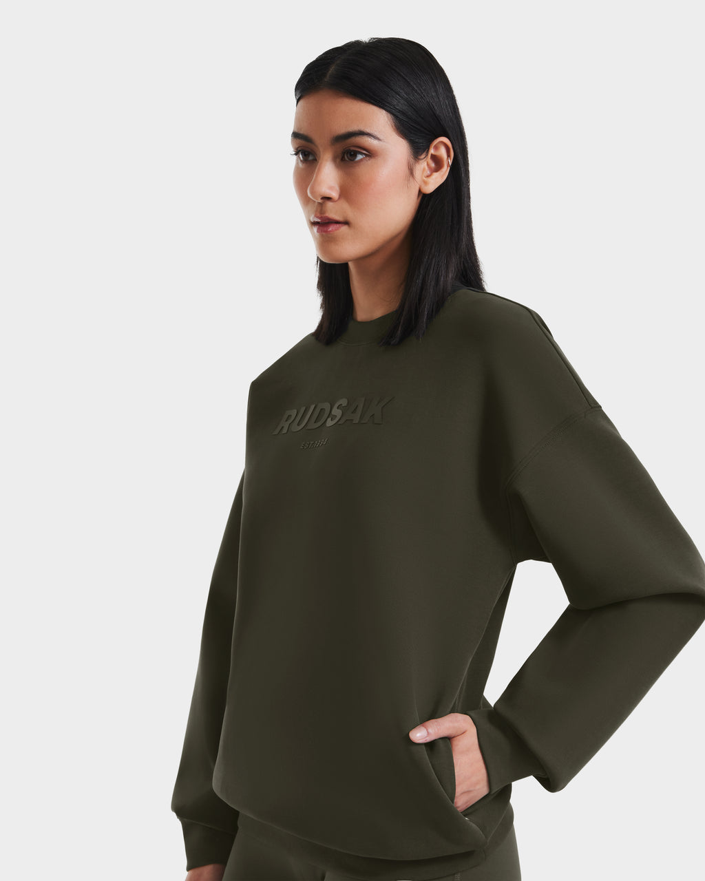 A woman with straight black hair wears the Rudsak BRYN - 9524007 Dark Olive W cotton crewneck sweatshirt, posing with one hand on her hip against a plain white background.