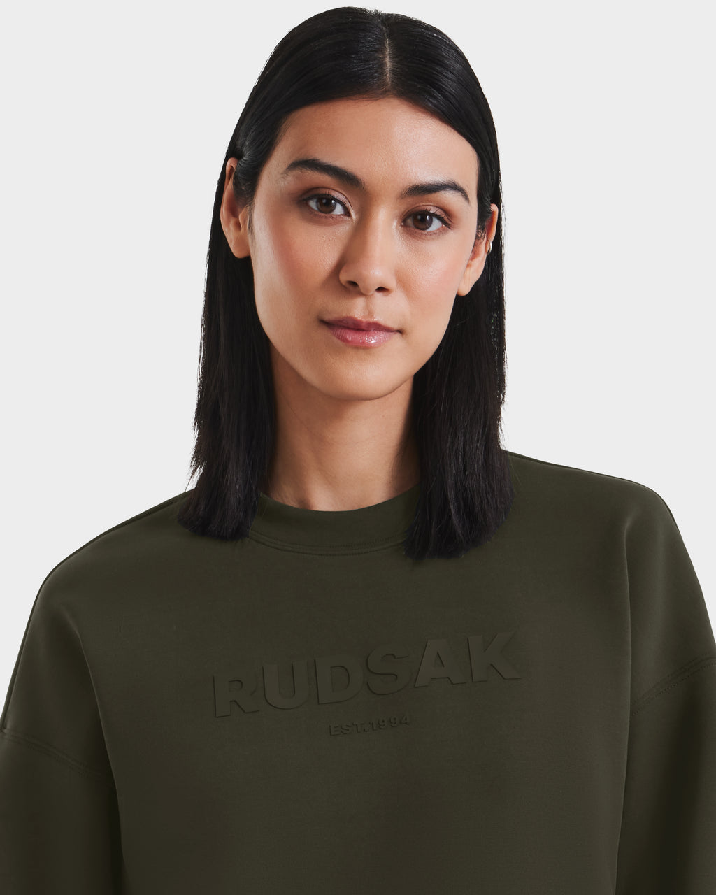 A woman with straight, shoulder-length dark hair models Rudsak’s BRYN - 9524007 Dark Olive W cotton crewneck sweatshirt with an embossed logo, standing against a plain white background.