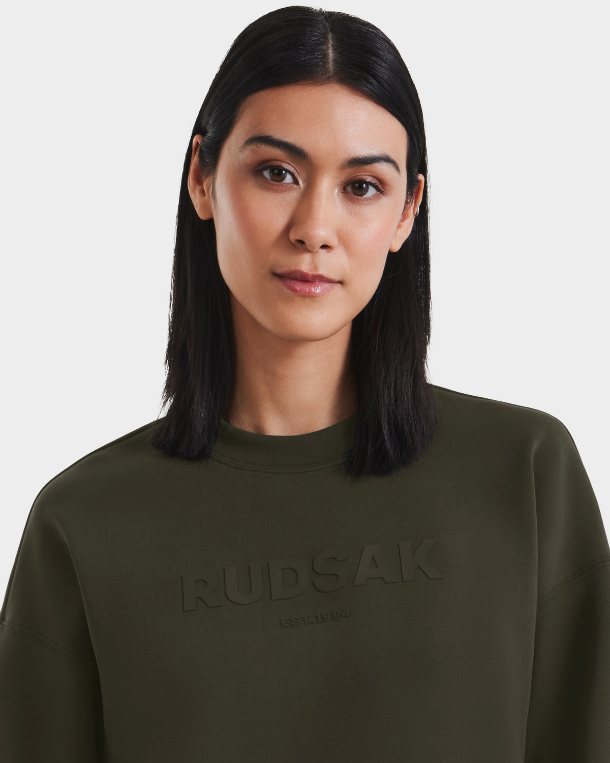 A woman with straight, shoulder-length dark hair models Rudsak’s BRYN - 9524007 Dark Olive W cotton crewneck sweatshirt with an embossed logo, standing against a plain white background.
