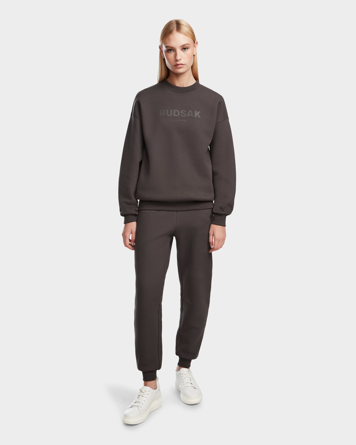 A woman with straight blonde hair models the Rudsak BRYN - 9524007 Grey W crewneck sweatshirt and jogger pants, paired with white sneakers, against a plain white background.