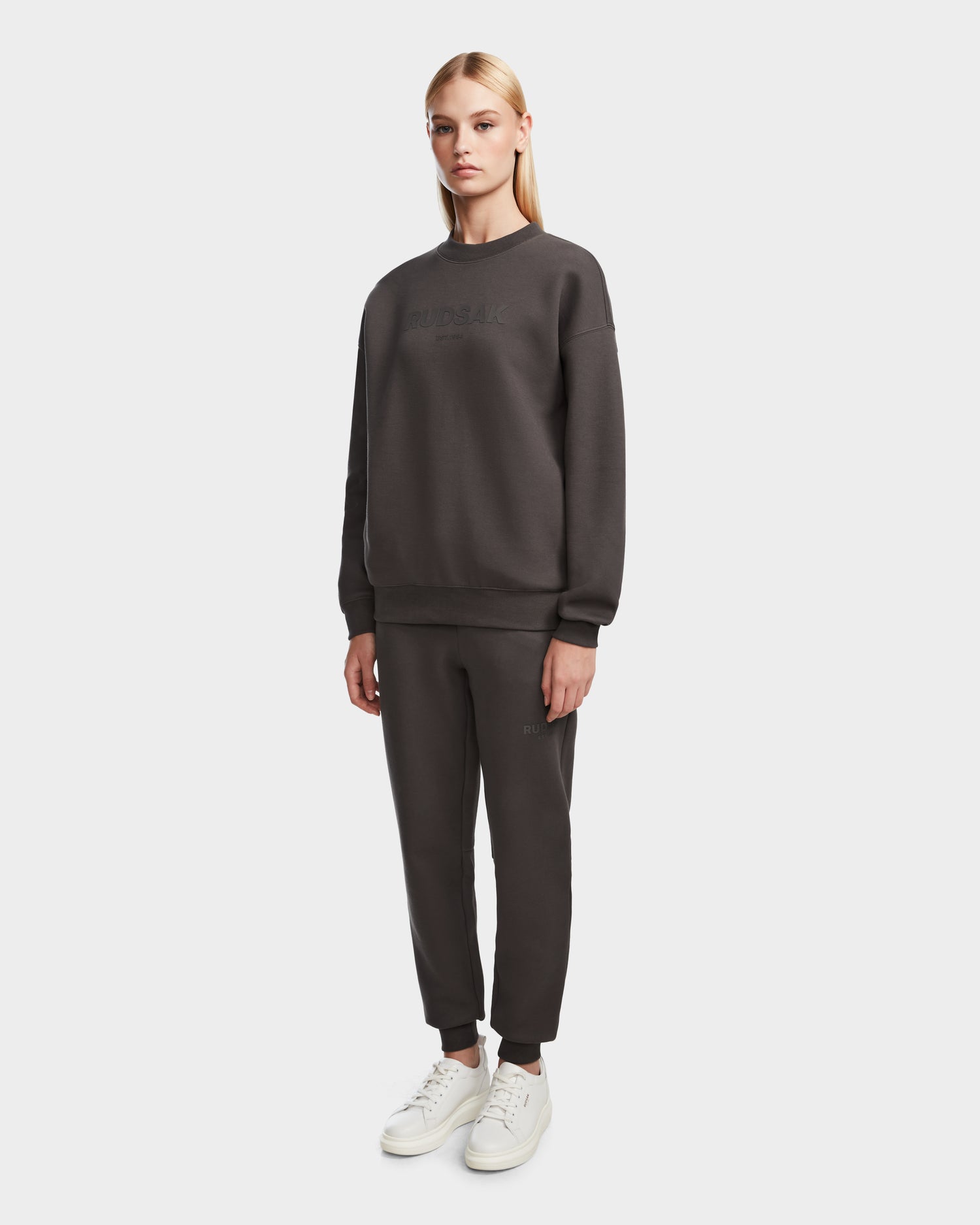 A woman with straight blonde hair stands against a plain white background wearing Rudsak’s BRYN - 9524007 Grey W crewneck sweatshirt and matching sweatpants with white sneakers, showcasing a timeless look.