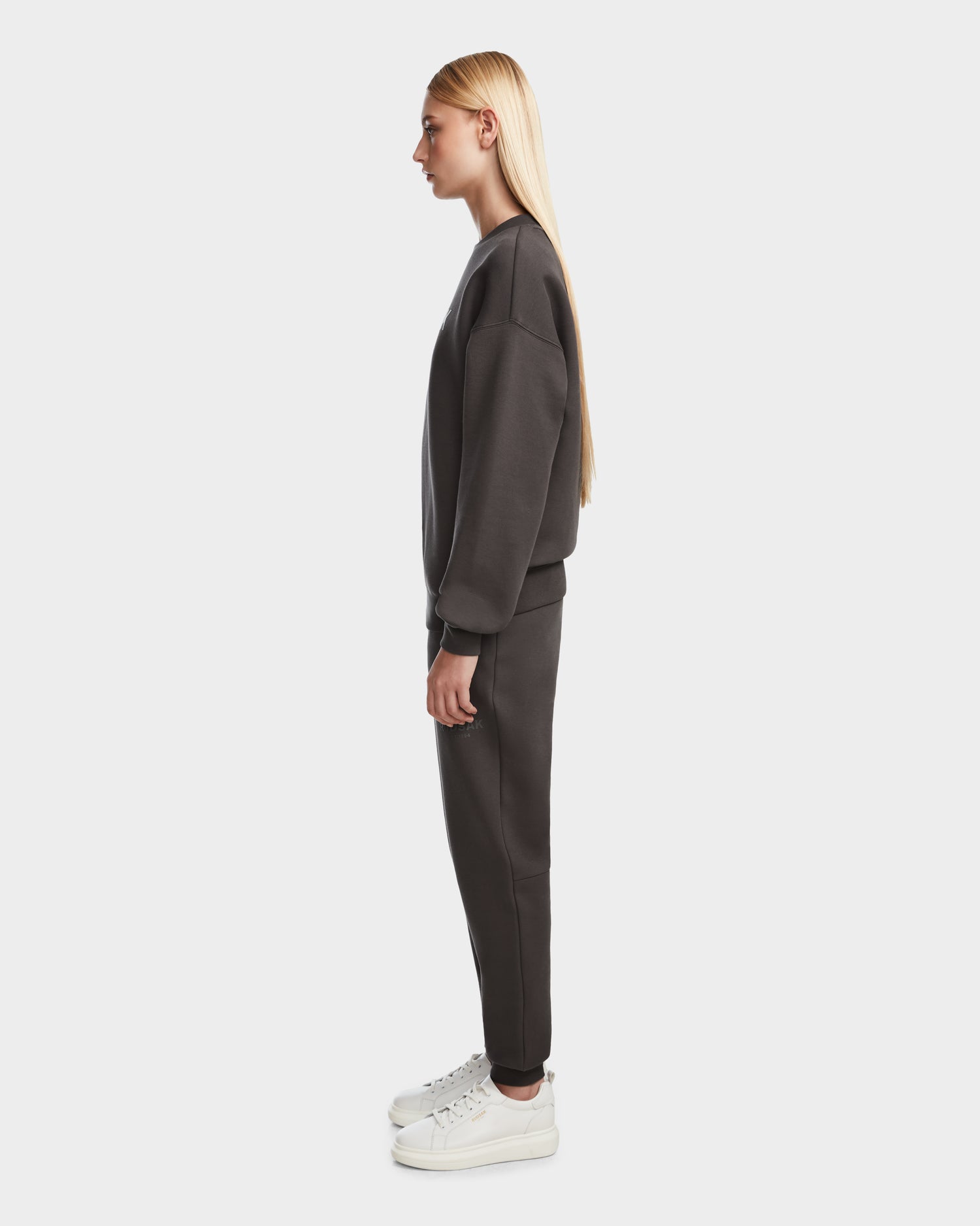 A woman in profile wears Rudsaks BRYN - 9524007 Grey W loose-fit, genderless cotton crewneck sweatshirt and matching jogger pants with white sneakers, set against a plain light background.