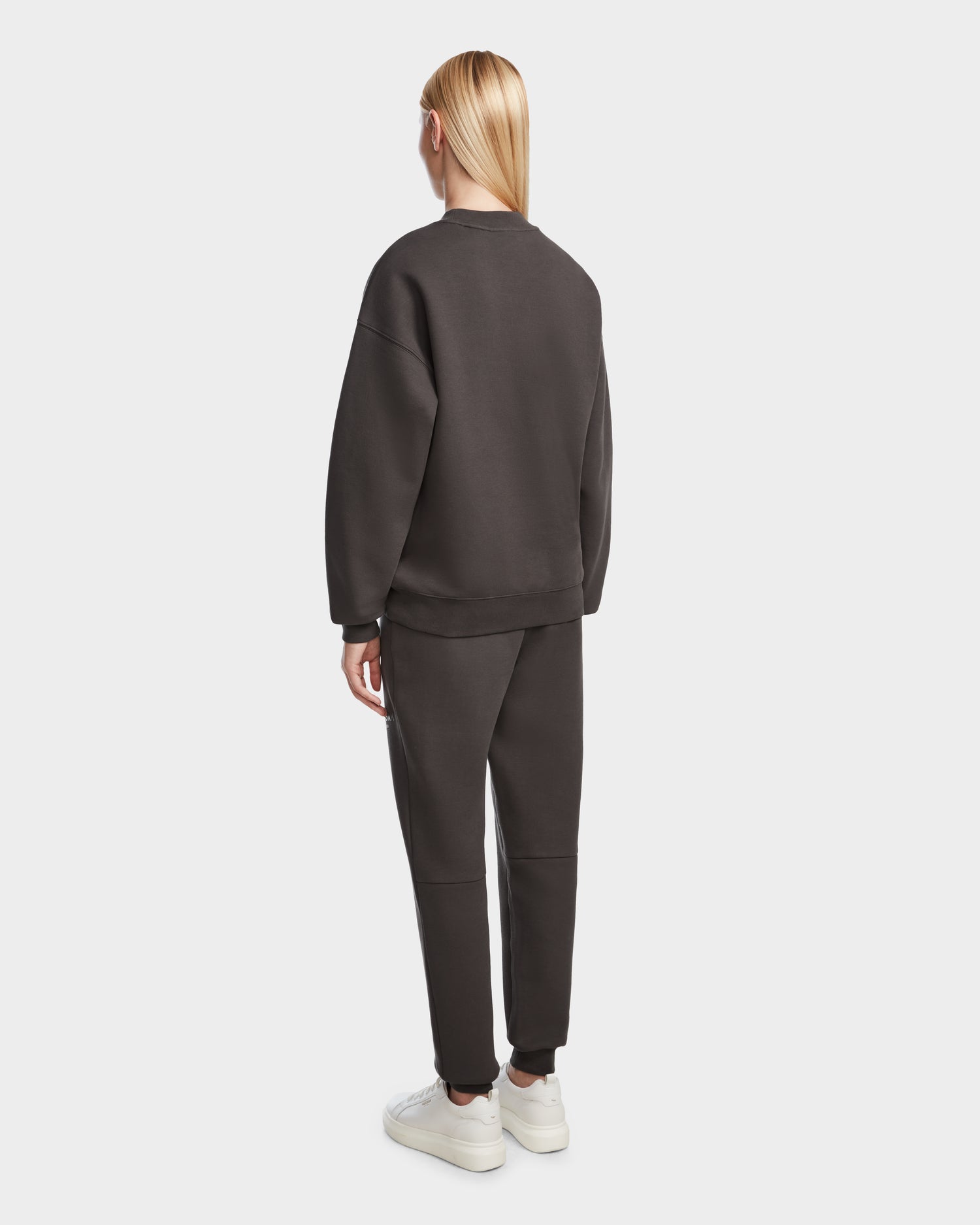 A person with straight blonde hair wears the Rudsak BRYN - 9524007 Grey W cotton crewneck sweatshirt and matching pants, paired with white sneakers. They stand facing away from the camera against a plain white background, showcasing timeless design.