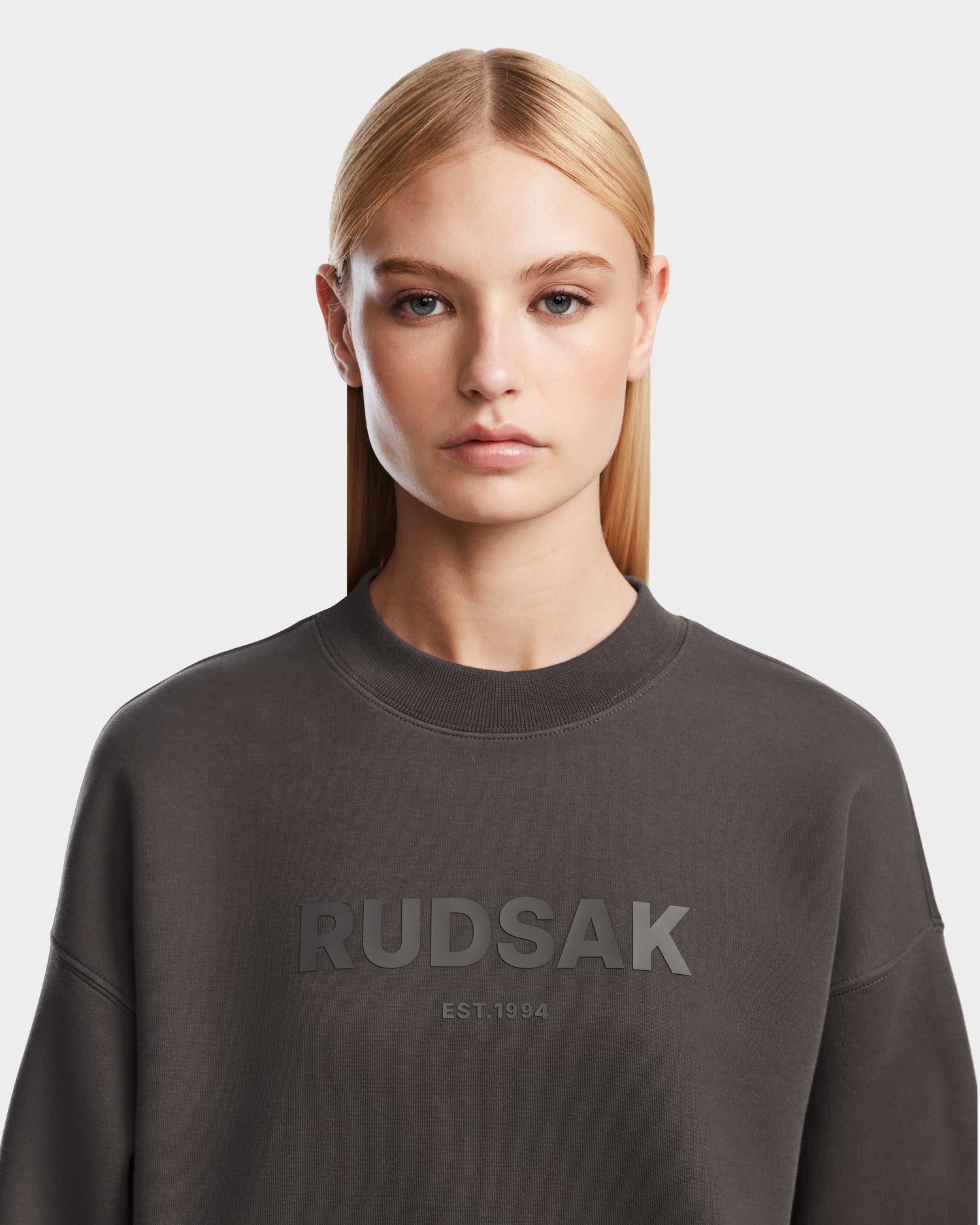A woman with straight blonde hair and a neutral expression wears the Rudsak BRYN - 9524007 Grey W crewneck sweatshirt, featuring RUDSAK EST.1994 on the front, against a plain light background, showcasing its timeless design.