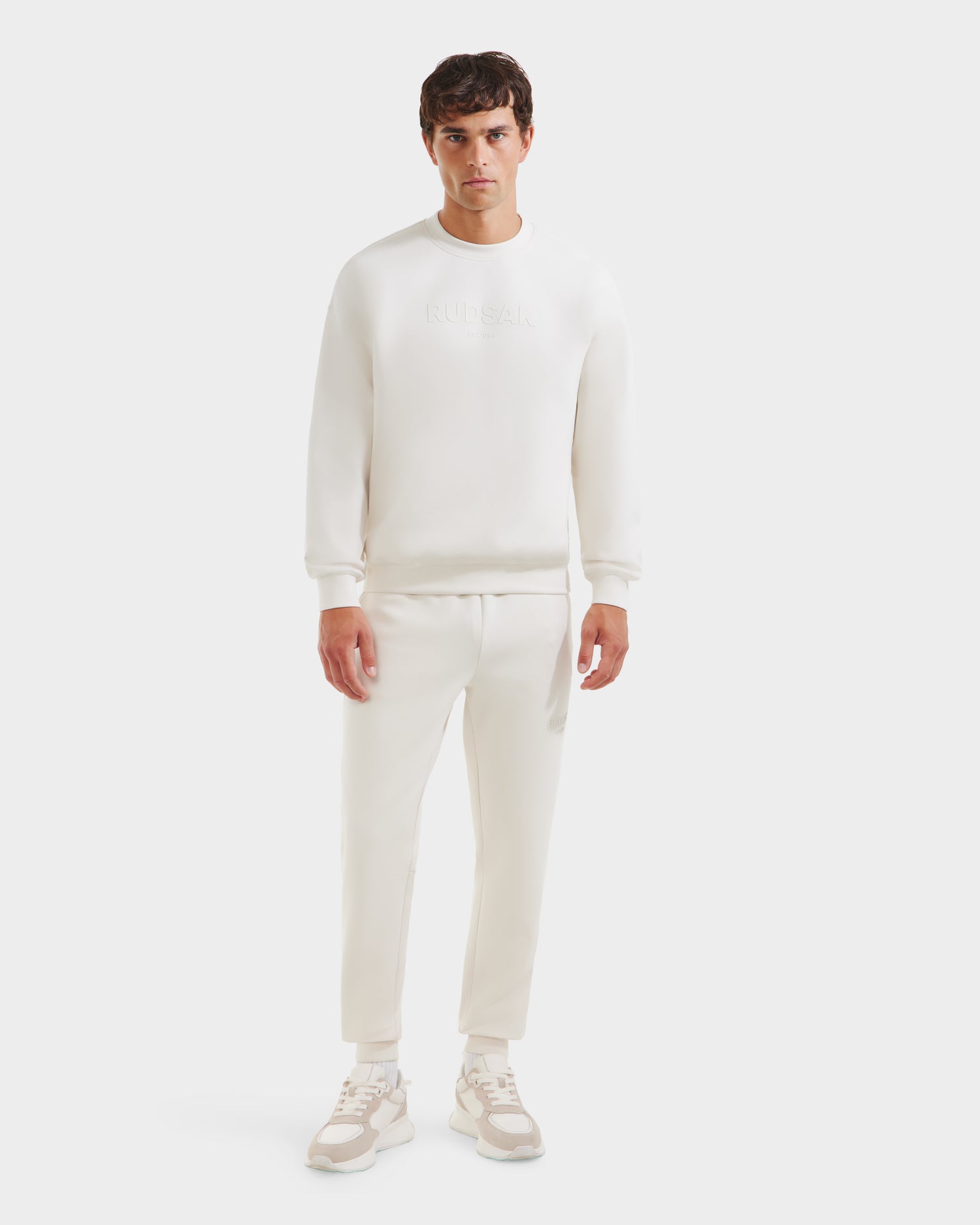 A man faces forward in a Rudsak BRYN - 9524007 Vanilla M crewneck sweatshirt with matching jogger pants and sneakers, presenting an everyday look against a plain white background.
