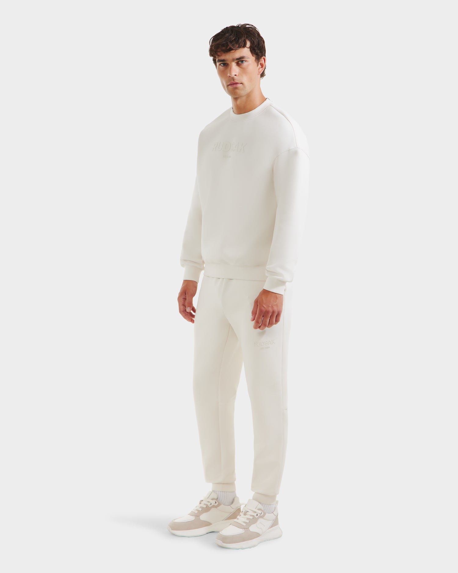 A man stands facing forward in a Rudsak BRYN - 9524007 Vanilla M cotton crewneck sweatshirt and joggers, paired with white sneakers with beige accents, all styled against a plain white background.