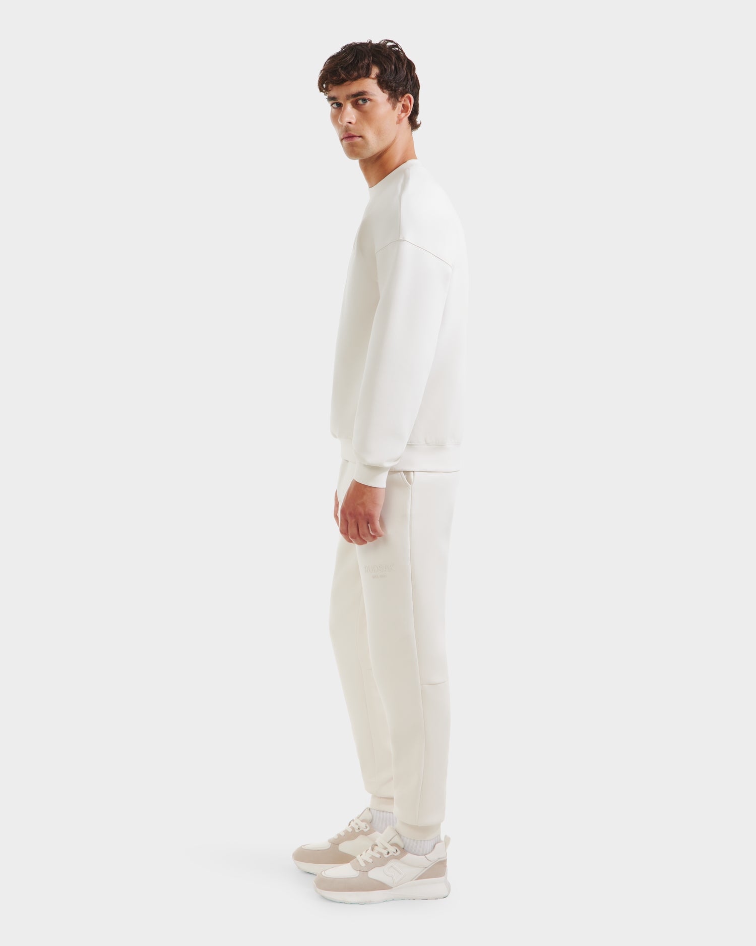 A man wears the Rudsak BRYN - 9524007 Vanilla M cotton crewneck sweatshirt and sweatpants with everyday fit and genderless sizing, paired with beige-and-white sneakers, standing sideways against a plain white background.