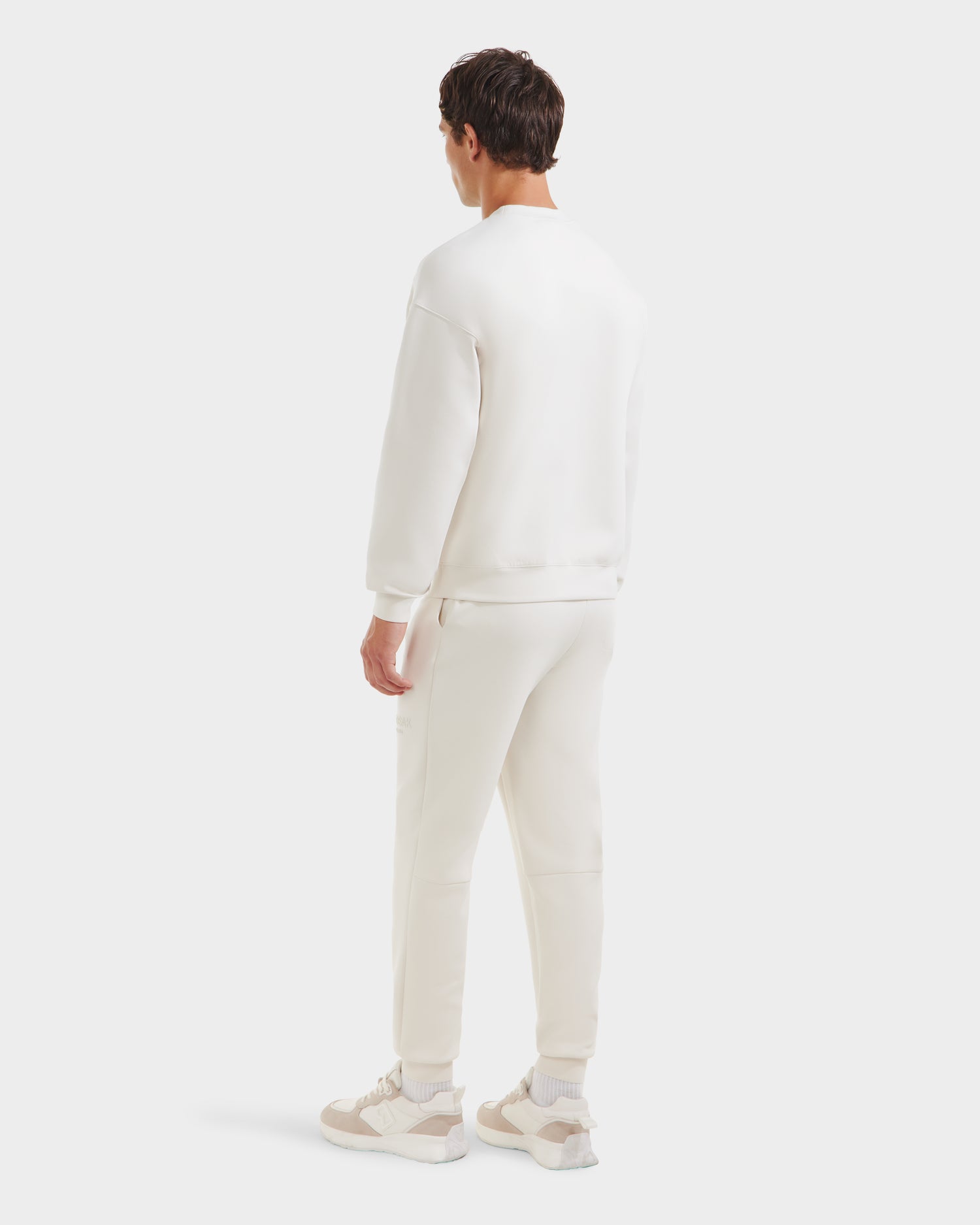 A person stands facing away, wearing the Rudsak BRYN - 9524007 Vanilla M cotton crewneck sweatshirt and matching joggers, paired with white sneakers with beige accents, against a plain light background.