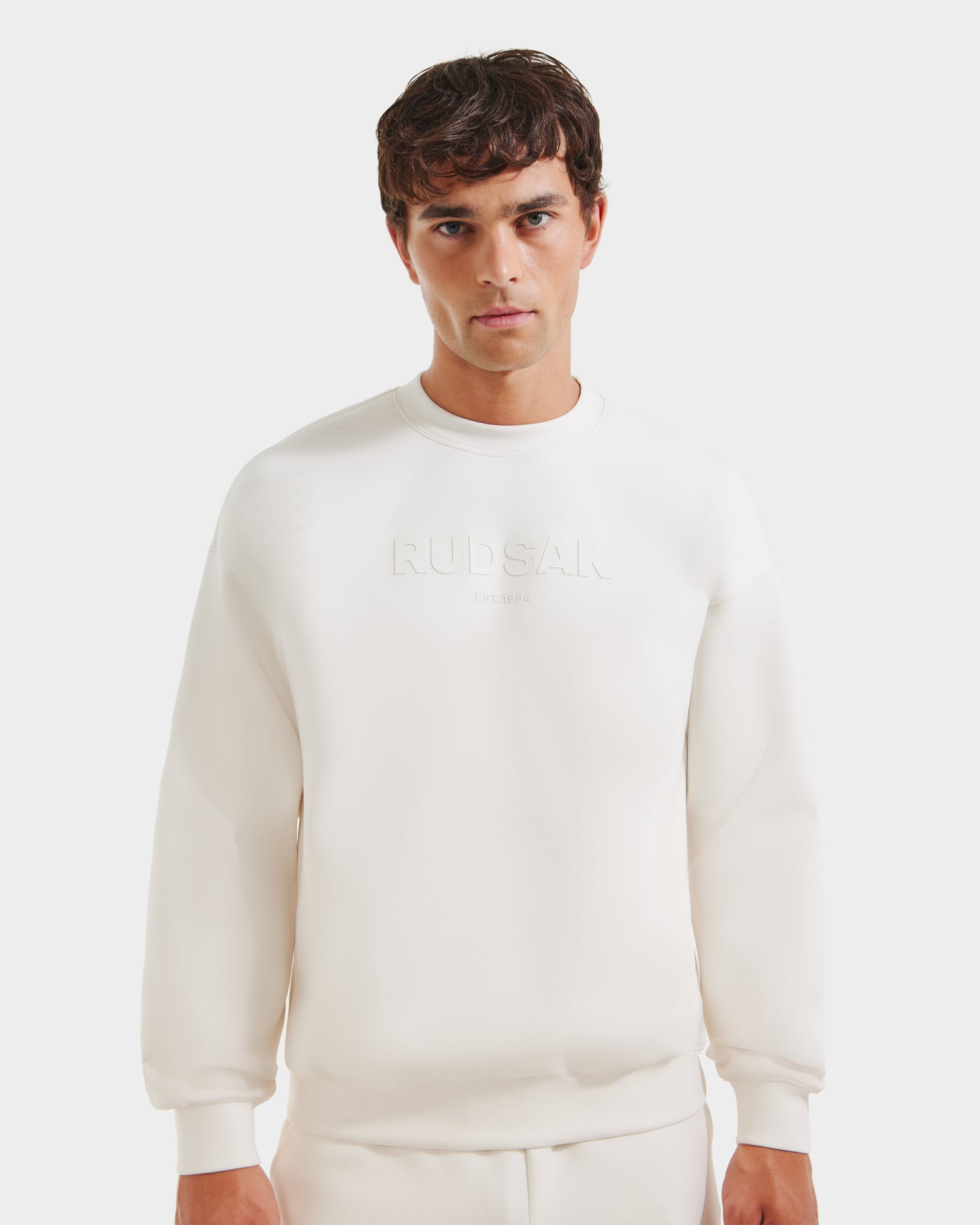 A man models the Rudsak BRYN - 9524007 Vanilla M, a white cotton crewneck sweatshirt with RUDSAK embossed on the chest, timeless design, and genderless sizing, standing against a plain white background.
