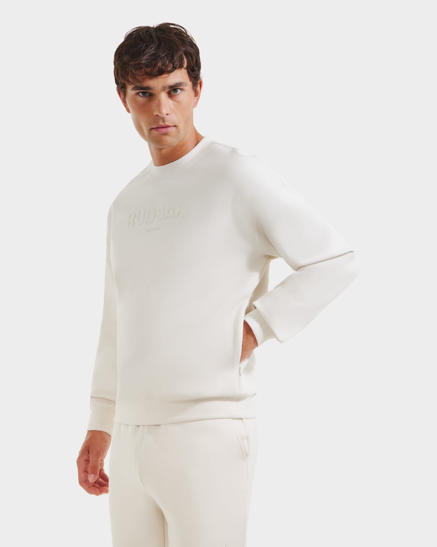A man models the Rudsak BRYN - 9524007 Vanilla M, an off-white cotton crewneck sweatshirt with subtle embossed chest text and genderless sizing, paired with matching pants against a plain white background.