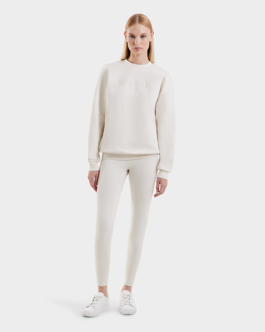 A woman faces forward in the Rudsak BRYN - 9524007 Vanilla W cotton crewneck sweatshirt with matching leggings and white sneakers, creating an everyday look against a plain white background.