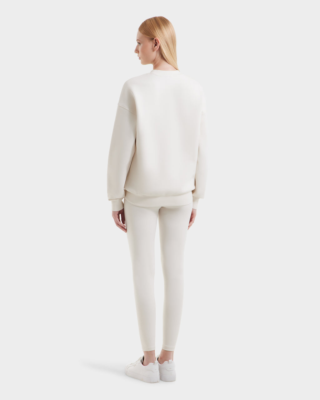 A woman with straight blonde hair faces away, wearing the Rudsak BRYN - 9524007 Vanilla W off-white crewneck sweatshirt and matching pants with white sneakers, showcasing a casual look against a plain light background.