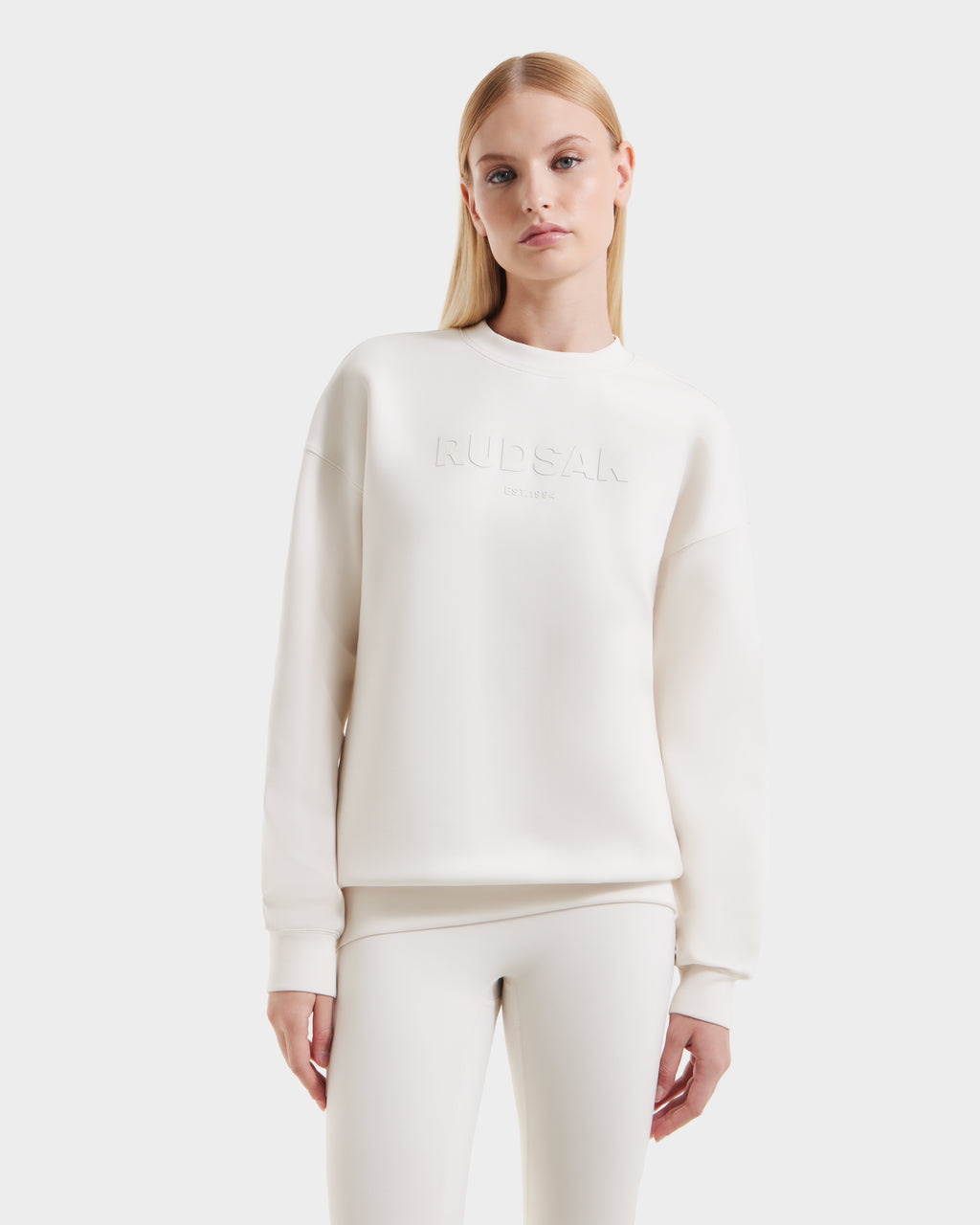 A person with long blonde hair models the Rudsak BRYN - 9524007 Vanilla W cotton crewneck sweatshirt, featuring embossed branding, styled with matching white pants against a plain white background.